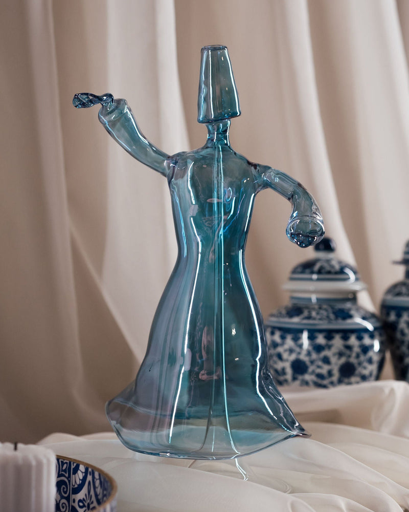 A hand-blown blue glass Darweesh, a type of traditional lantern, with glass support, displayed in a setting with white curtains and decorative blue and white vases in the background.